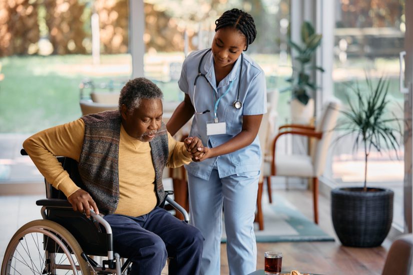 African American senior man getting up from his wheelchair with help of young nurse at residential care home.